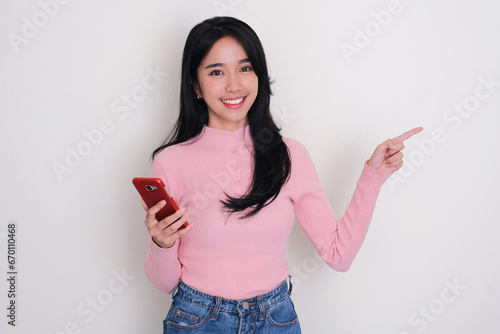 Young Asian woman smiling and pointing to the left side while holding mobile phone photo