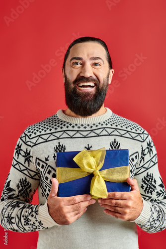 pleased bearded man in winter sweater with ornament holding Christmas present on red background © LIGHTFIELD STUDIOS