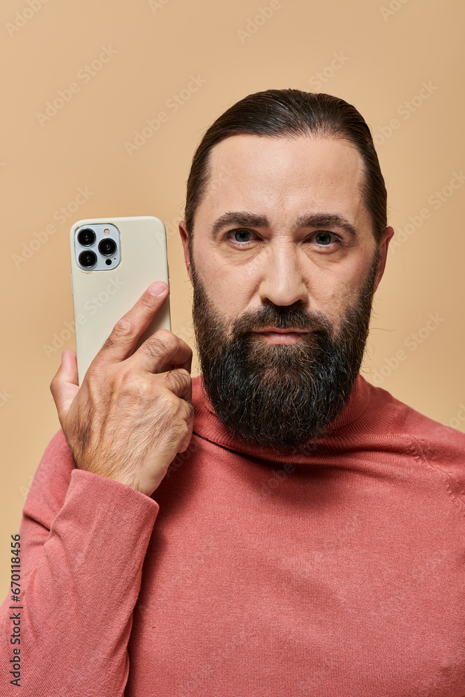 portrait of good looking bearded man in turtleneck jumper holding smartphone on beige background