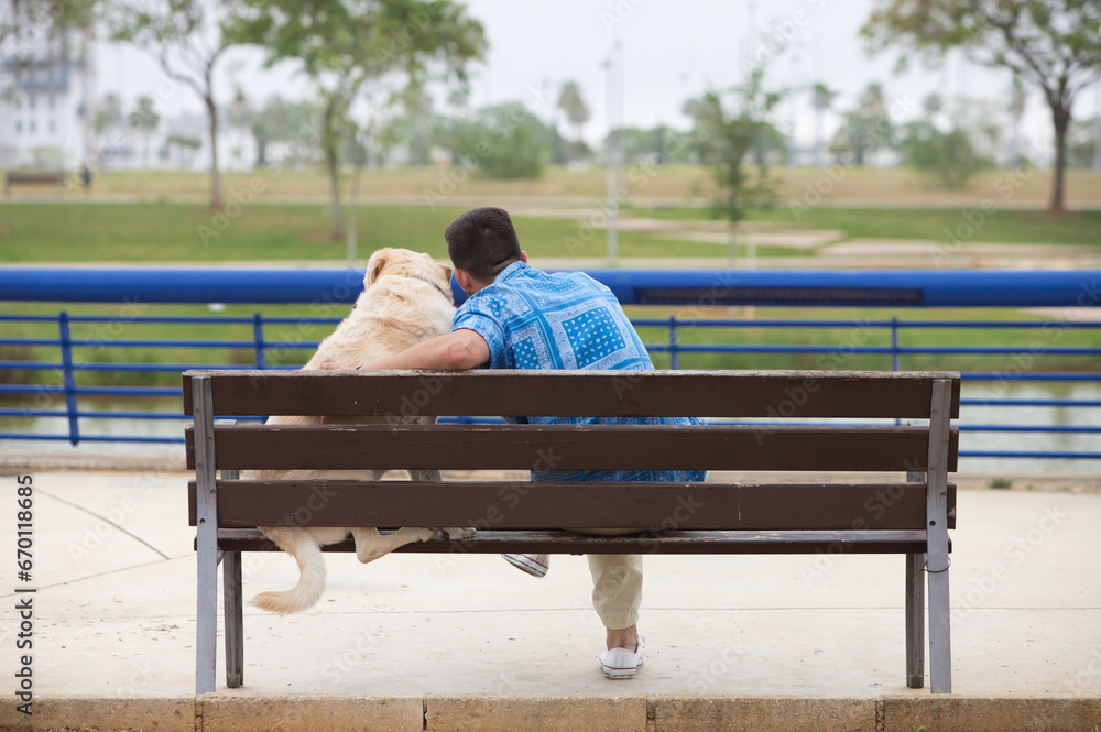 Handsome young man and his Labrador retriever dog sitting on a wooden bench in the park. The man and his pet have their heads together. The picture is taken from behind. Concept pets and animals. Handsome young man and his Labrador retriever dog sitting on a wooden bench in the park. The man and his pet have their heads together. The picture is taken from behind. Concept pets and animals.