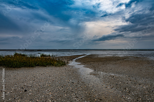 Ein Rinnsal fließt zwischen Muscheln und Wolken in Richtung Wattenmeer wo trotz Ebbe einiges an Wasser steht