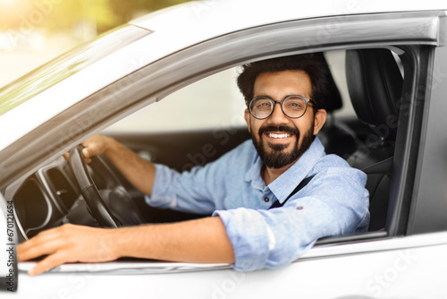 Cheerful millennial indian guy with glasses driving white auto