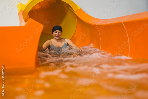 Amused laughing senior adult woman in striped swimsuit and black hair cap sliding on an orange indoor water slide. High quality photo photo