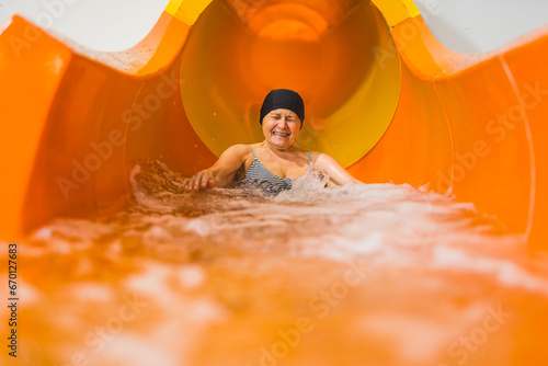 Laughing amused elderly caucasian female pensioner in black hair cap sliding on an orange water slide. High quality photo