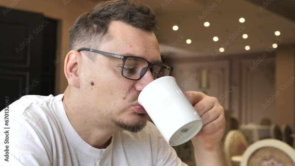 Happy young man drinking a hot drink from a mug. A satisfied smiling guy drinks tea while sitting at a table in a restaurant. Man indoors enjoying a cup of coffee or tea Happy young man drinking a hot drink from a mug. A satisfied smiling guy drinks tea while sitting at a table in a restaurant. Man indoors enjoying a cup of coffee or tea