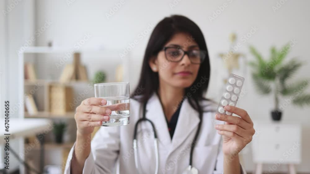 Friendly female therapist in glasses showing white jar of medicine while sitting at desk. Experienced indian woman in white coat recommending best alternative of treating at doctor's office. Friendly female therapist in glasses showing white jar of medicine while sitting at desk. Experienced indian woman in white coat recommending best alternative of treating at doctor's office.