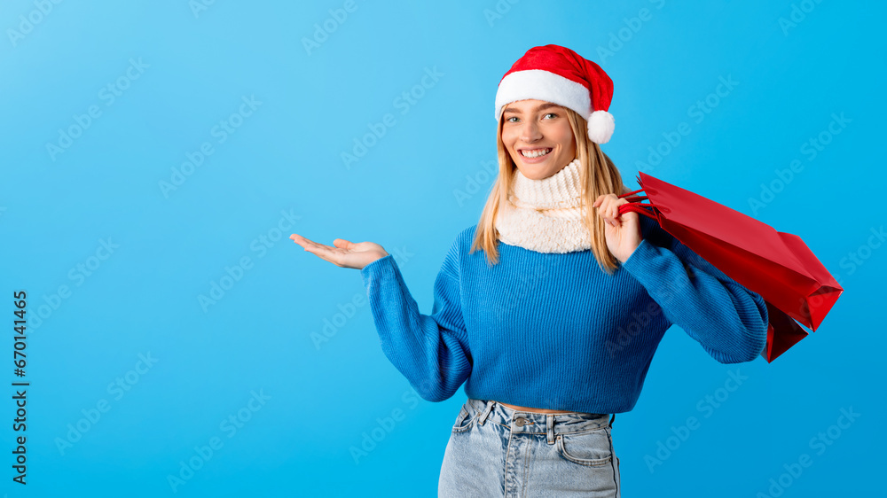 Obraz premium Joyful woman in Santa hat with shopping bag showing something invisible, blue backdrop
