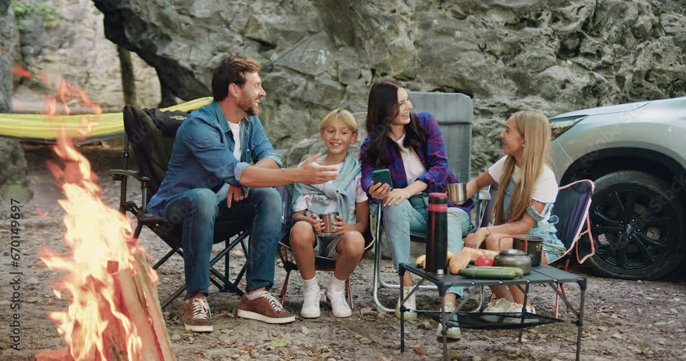 Happy young handsome parents and their children using smartphone watching video or browsing internet together online sitting near campfire during weekend outdoors in nature. Leisure and technology Happy young handsome parents and their children using smartphone watching video or browsing internet together online sitting near campfire during weekend outdoors in nature. Leisure and technology