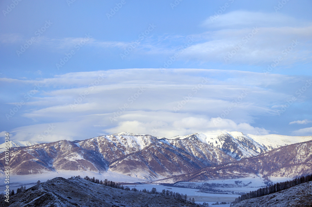 Cold snowy mountain landscape at sunset. Lenticular clouds. Panoramic view of snowy mountain peaks and slopes of North Chuyskiy ridge at sunset. Russia, Siberia, Altai mountains.