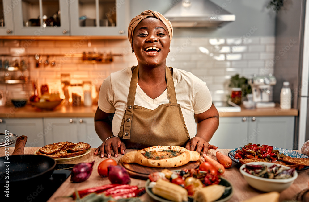 Domestic culinary. Cheerful african american woman in turban and apron standing behind kitchen table full of delicious traditional meals. Plus size housewife cooking at cozy home for beloved family.