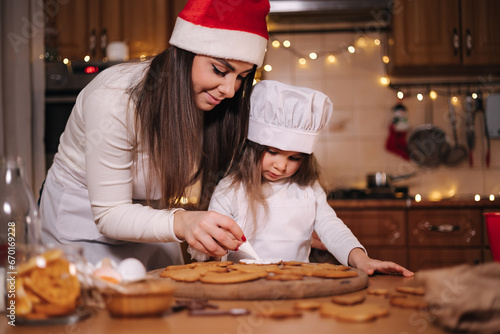 Happy little girl with her beautiful mom make gingerbread at home. Christmas decoration at kitchen. Fir tree with fairy lights