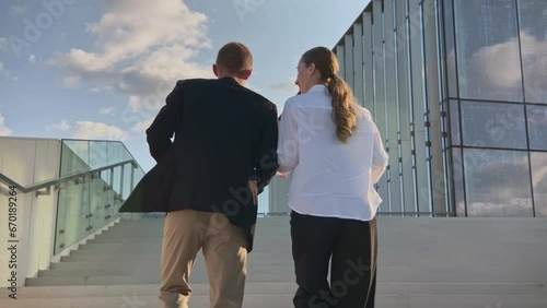 Two caucasian business partners man and woman walk together up stairs near office glass building, colleagues talking about work professional tasks spending time during break. Back view, slow motion
 photo