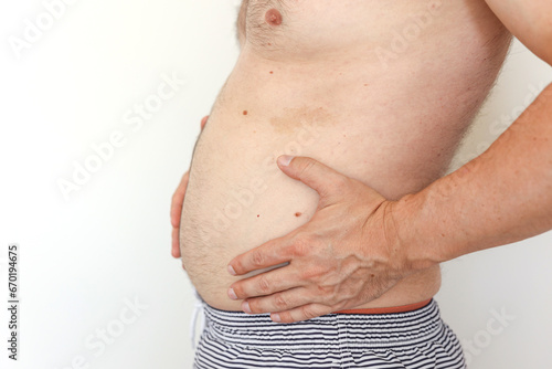 Cropped photo of man is touching his naked not fit belly waistline with loose skin suffering from extra weight. Concept of unhealthy eating and lifestyle, health problems of obese people.