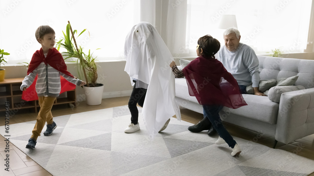 Fototapeta premium Aged great-grandfather spend time with three great-grandsons at home, kids in festive Halloween costumes running in living room, having fun together, enjoy active game, friendship and family pastime