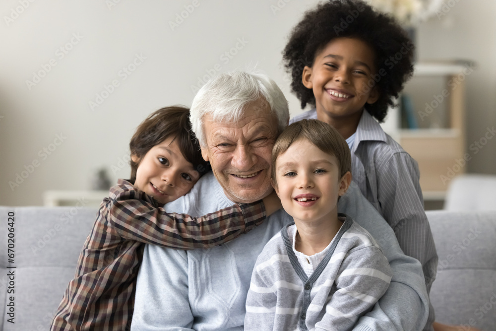 Three multiracial grandsons hug their hoary great-grandfather, smile look at camera, sitting on sofa posing together for family photo. Multigenerational family bond, ties, unity. Offspring portrait Three multiracial grandsons hug their hoary great-grandfather, smile look at camera, sitting on sofa posing together for family photo. Multigenerational family bond, ties, unity. Offspring portrait