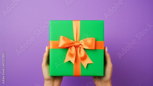 Anonymous person showing bright green carton gift box with ribbon and purple bow on orange table against violet background