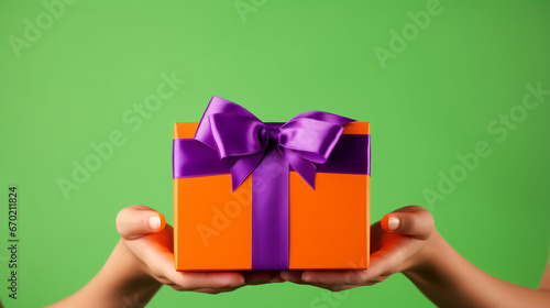 Anonymous person showing bright orange carton gift box with ribbon and purple bow against green background