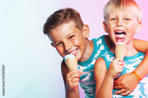 Tasty treat concept. Happy two young handsome brothers wearing sleeveless shirts with sharks  hugging  eating melting ice cream in waffle cone over pink and blue background. Copy-space