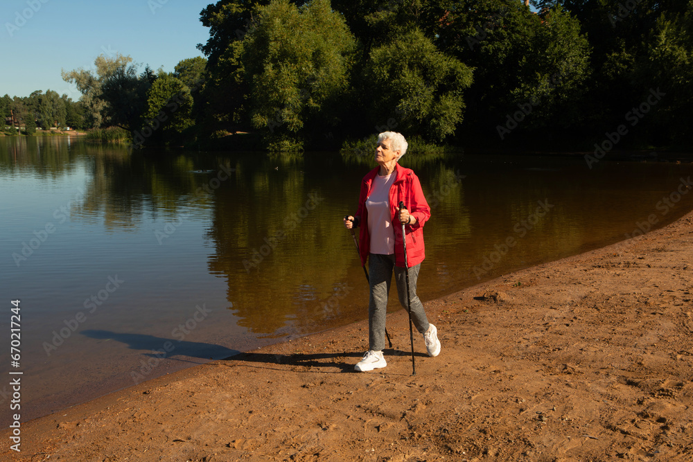 Grey-haired woman walking with tracking sticks on the beach near lake Grey-haired woman walking with tracking sticks on the beach near lake