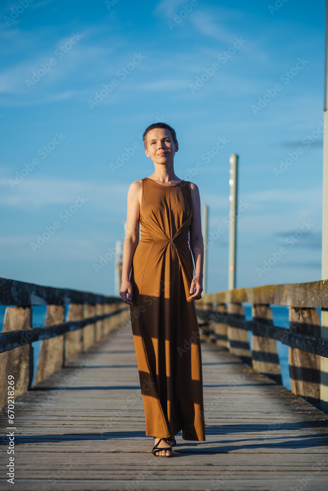 Women's independence, a woman with a short haircut in a long dress walks along the pier against the blue sky.