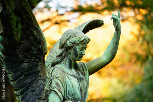 an angel with wings in a cemetery in front of luminous autumn leaves in the background raises one arm to the sky photo