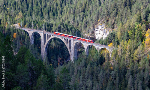 A red passenger train is crossing the famous Wiesener viaduct on the train line Davos - Filisur, the highest viaduct in swiss alps photo