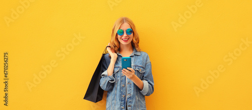 Beautiful happy young woman looking at mobile phone with shopping bag wearing denim jacket on yellow studio background