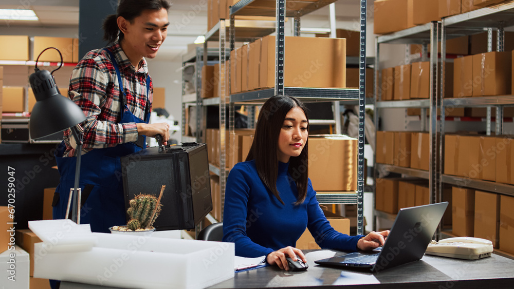 Fototapeta premium Asian team working on product inventory in warehouse, checking merchandise before shipment and distribution. Young woman doing quality control at desk, small business development.