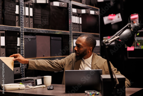 Detective analyzing crime case and searching police report while conducting investigation. African american policeman working in office and studying archival records in evening