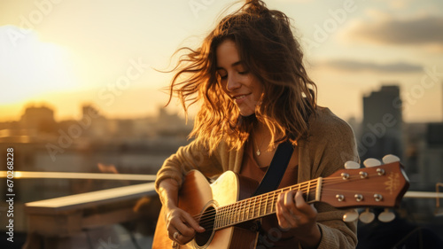 Young woman plays guitar on roof terrace at sunset, adult girl guitarist practices music. Female player with acoustic instrument outside home. Concept of summer, city, musician