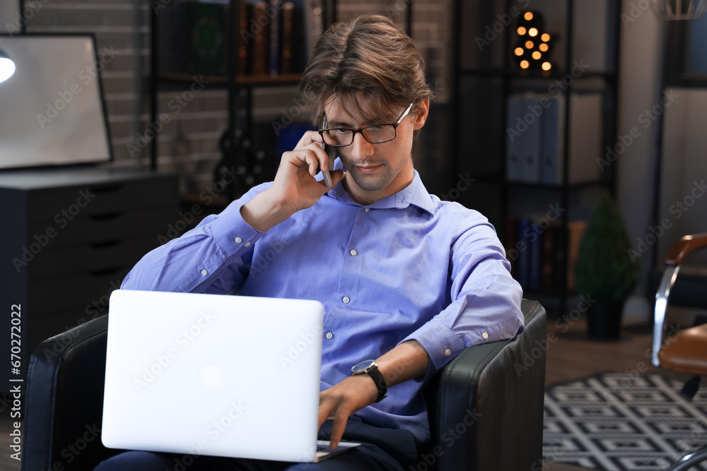 Young businessman sitting on armchair in office, talking by mobile phone and working with laptop Young businessman sitting on armchair in office, talking by mobile phone and working with laptop