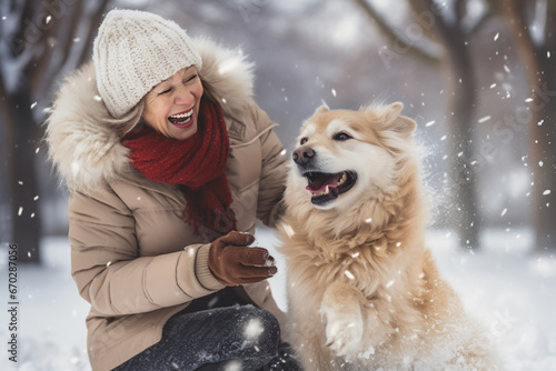 A senior caucasian woman is playing happily with the dog in the snow with a winter coat and winter hat in a in snow covered country landscape during day in winter while snowing