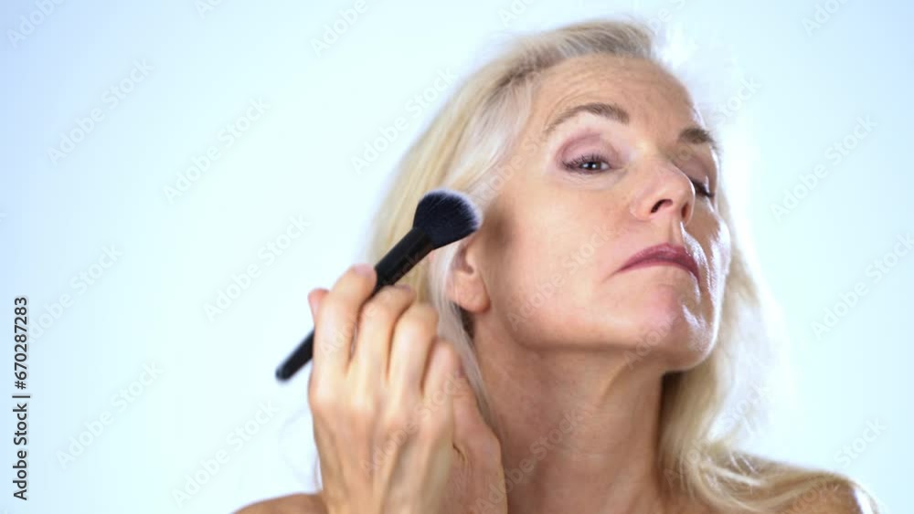 Closeup portrait of 50s, 60s, mature middle aged nude woman applying natural concealer makeup powder foundation with large brush on clean face skin smoothing wrinkles, white background. Closeup portrait of 50s, 60s, mature middle aged nude woman applying natural concealer makeup powder foundation with large brush on clean face skin smoothing wrinkles, white background.