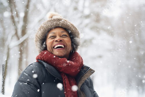 A senior african american woman is playing in the snow happily with a winter coat and a winter hat in a in snow covered forest during day in winter while snowing