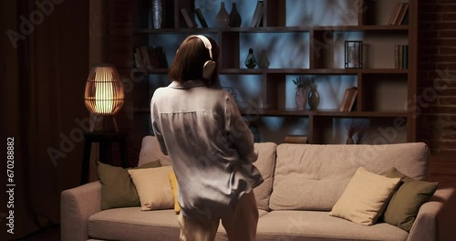 Joyful Caucasian girl dances freely in the living room, wearing headphones that transport her into the rhythm of the music. Her face lights up with happiness, and body moves gracefully to the beat.