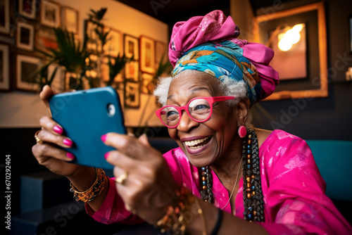 A senior african american woman is is making a selfie while smiling with a telephone behind a computer while gaming a low tech social media woman