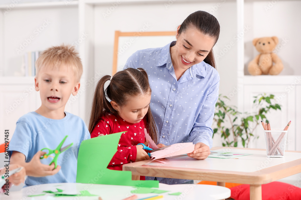 Nursery teacher and cute little children making toys from color paper at desks in kindergarten. Playtime activities