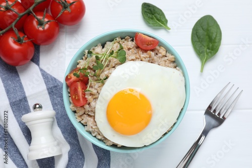Tasty boiled oatmeal with fried egg, tomato and microgreens served on white wooden table, flat lay