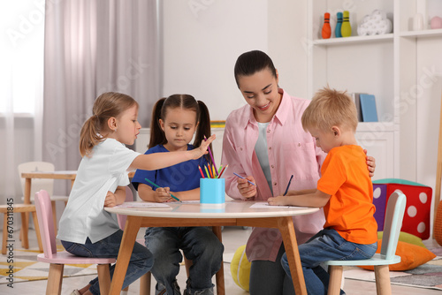 Nursery teacher and group of cute little children drawing at desk in kindergarten. Playtime activities