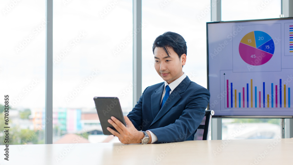 Asian professional successful male businessman employee in formal business suit sitting working using tablet computer in company office meeting room with graphs charts on big computer monitor screen Asian professional successful male businessman employee in formal business suit sitting working using tablet computer in company office meeting room with graphs charts on big computer monitor screen