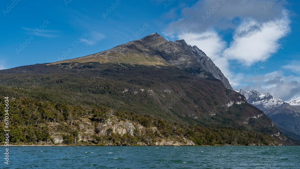 A picturesque mountain on a background of blue sky and clouds. The forest grows on rocky slopes. In the foreground is an emerald lake with ripples and foam flaps on the surface. Lago Roca. Argentina. 