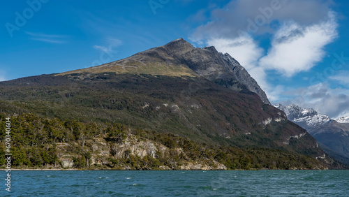 A picturesque mountain on a background of blue sky and clouds. The forest grows on rocky slopes. In the foreground is an emerald lake with ripples and foam flaps on the surface. Lago Roca. Argentina. 