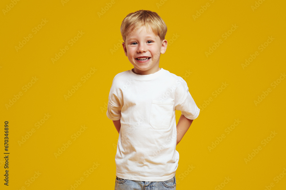 Positive blonde boy in casual white t-shirt keeping hands behind back and smiling for camera while standing against yellow background. Positive blonde boy in casual white t-shirt keeping hands behind back and smiling for camera while standing against yellow background.