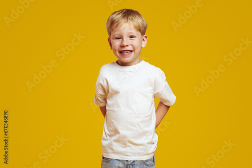 Positive blonde boy in casual white t-shirt keeping hands behind back and smiling for camera while standing against yellow background.