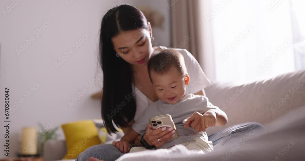 Mom and child on sofa looking to smartphone. Cinematic Chinese, Japanese, Korean, Asian. Beauty and tenderness of mother's embrace, showcasing the emotional connection between mother and beloved child Mom and child on sofa looking to smartphone. Cinematic Chinese, Japanese, Korean, Asian. Beauty and tenderness of mother's embrace, showcasing the emotional connection between mother and beloved child