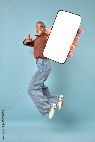 Young excited happy African American woman jumping up and showing off mobile phone and pointing finger at white screen of gadget with copy space for advertising application located in turquoise studio