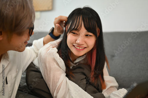 Happy young Asian woman lying on couch and watching movie on digital tablet with boyfriend at home