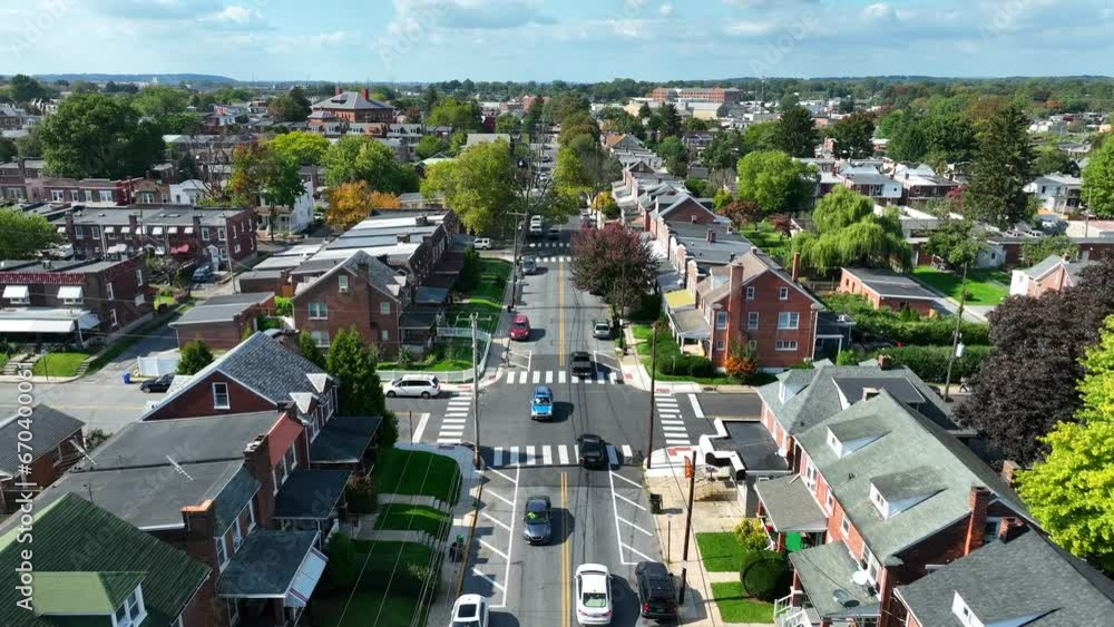 Aerial view of a suburban neighborhood with brick houses, tree-lined streets, and cars driving along a main road on a clear day. American suburb in early autumn. Aerial view of a suburban neighborhood with brick houses, tree-lined streets, and cars driving along a main road on a clear day. American suburb in early autumn.