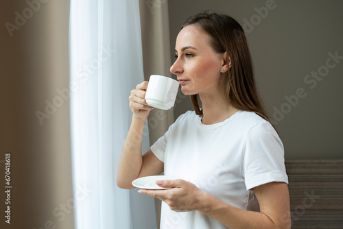 Young beautiful woman is looking through the window and drinking coffee in the morning. 