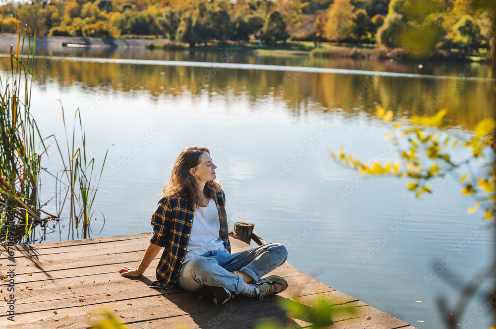 Young woman relaxing sitting on a wooden pier on a pond enjoying autumn nature